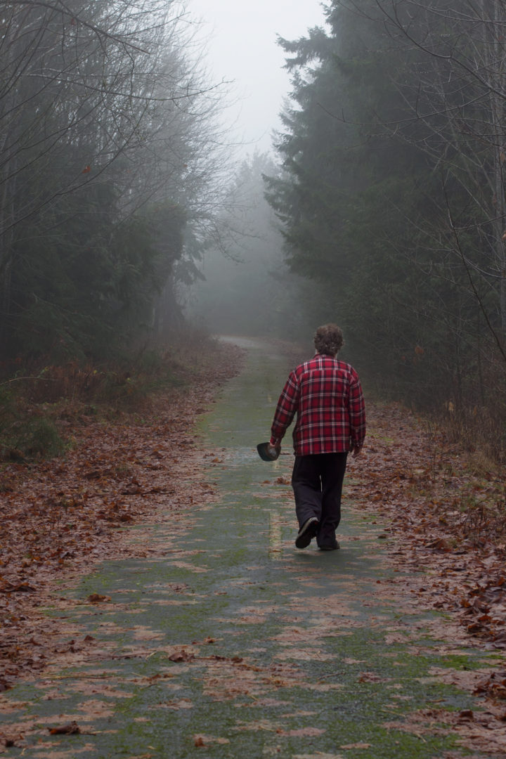 road covered in fall leaves with misty trees. man in plaid shirt walks away