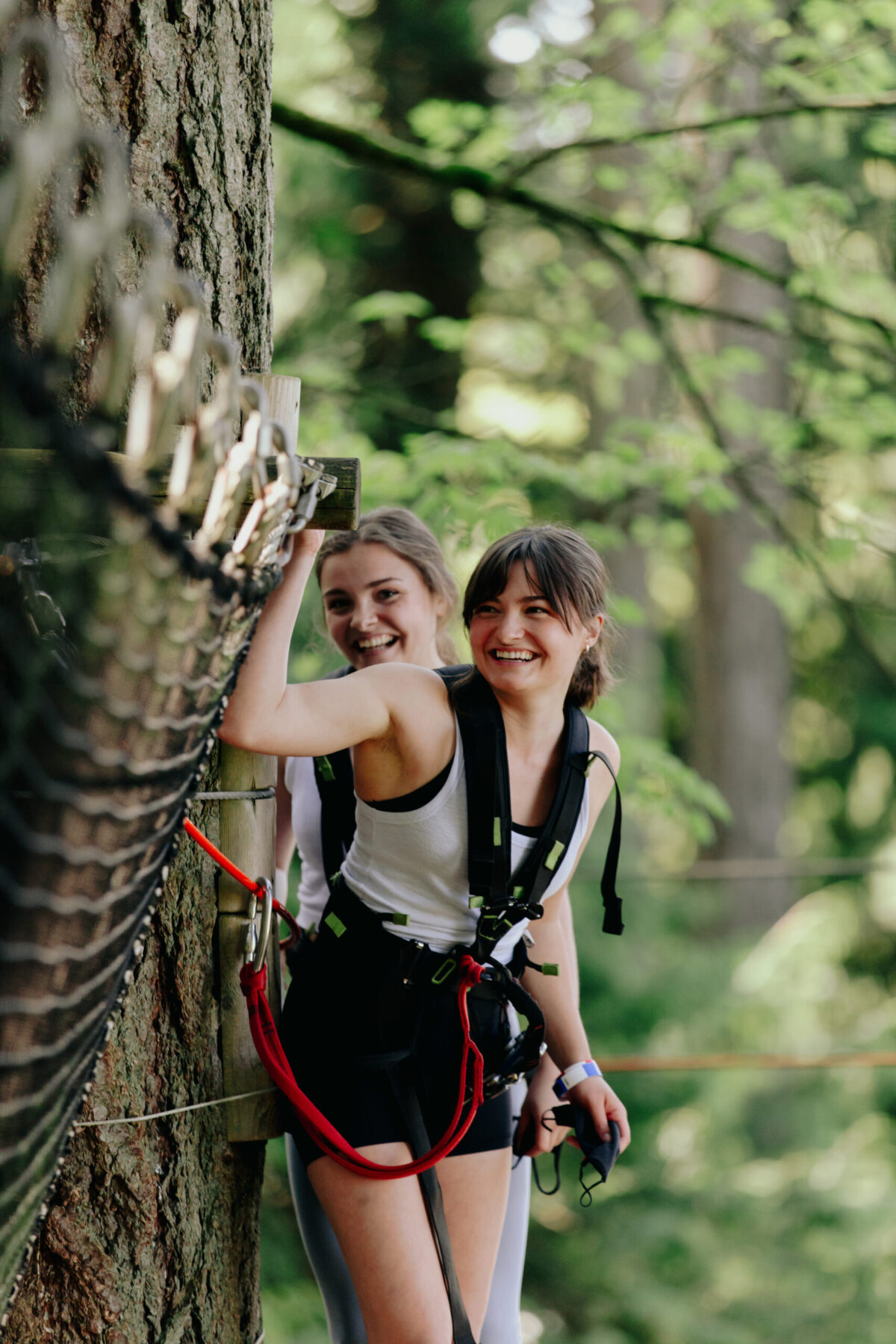 Two young women wearing harnesses stand on a wooden platform in a forest adventure course, smiling and holding onto a safety cable near a tree.