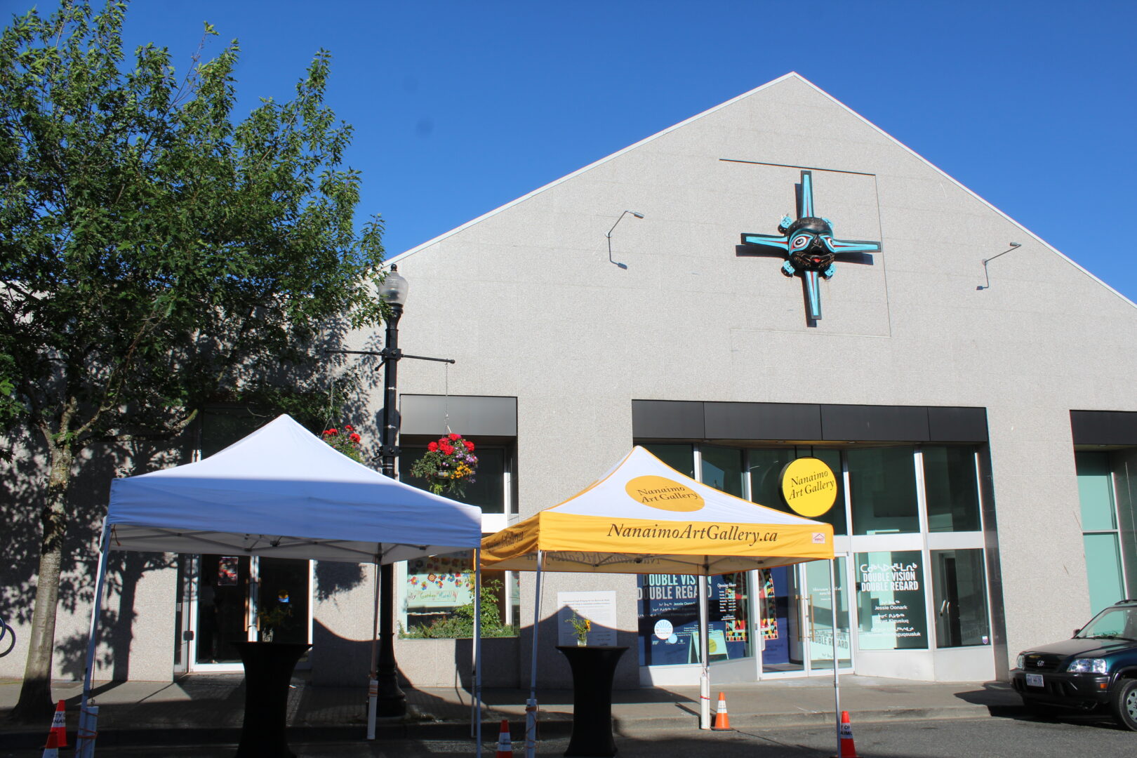 Two canopy tents, one white and one yellow, are set up in front of a modern building with large windows and a turquoise wall sculpture.
